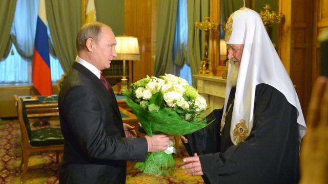 Putin offers flowers to Patriarch Kirill. Credits.