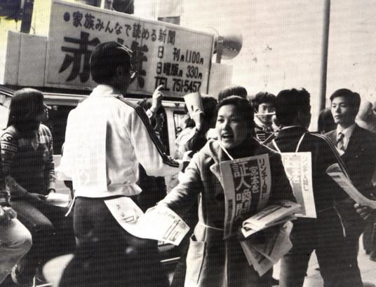 A young VOC activist handing out leaflets criticizing Chairman Miyamoto in front of a Communist Party campaign car.